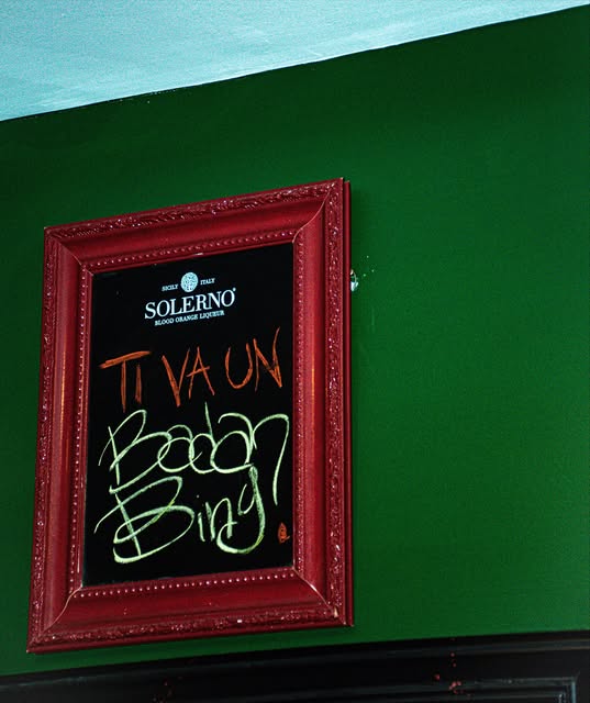 Lavagna in una cornice barocca rossa sulla parete verde: «Ti va un Bada Bing?» scritto col gesso